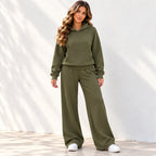 A woman modeling an olive green two-piece loungewear set consisting of a pullover hoodie and matching wide-leg sweatpants, paired with white sneakers against a white wall with soft leaf shadows.