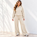 A woman modeling an beige two-piece loungewear set consisting of a pullover hoodie and matching wide-leg sweatpants, paired with white sneakers against a white wall with soft leaf shadows.