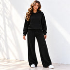 A woman modeling an black two-piece loungewear set consisting of a pullover hoodie and matching wide-leg sweatpants, paired with white sneakers against a white wall with soft leaf shadows.