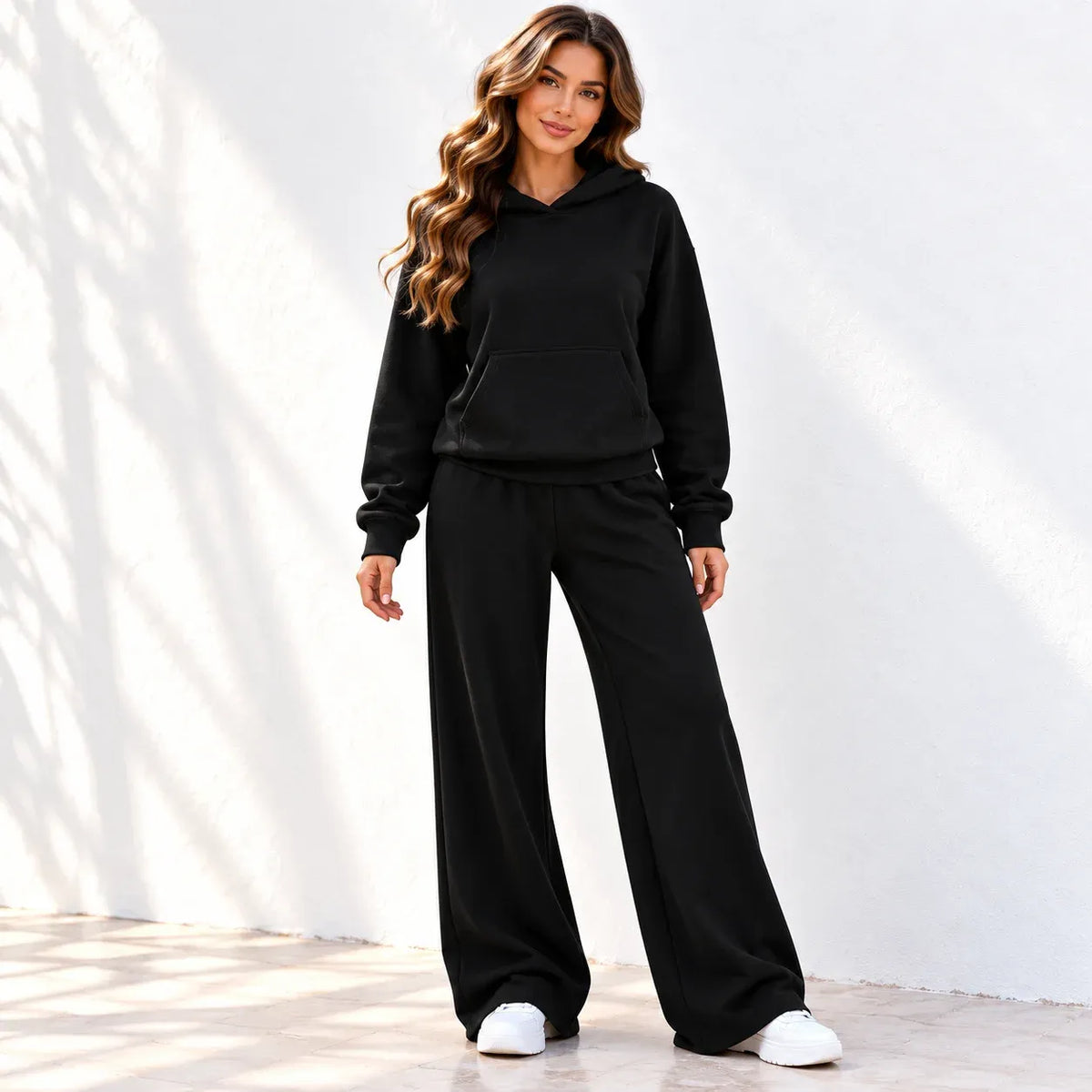 A woman modeling an black two-piece loungewear set consisting of a pullover hoodie and matching wide-leg sweatpants, paired with white sneakers against a white wall with soft leaf shadows.