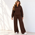 A woman modeling a matching brown two-piece loungewear set consisting of a pullover hoodie and wide-leg sweatpants, paired with white sneakers, standing against a white wall with soft leaf shadows.