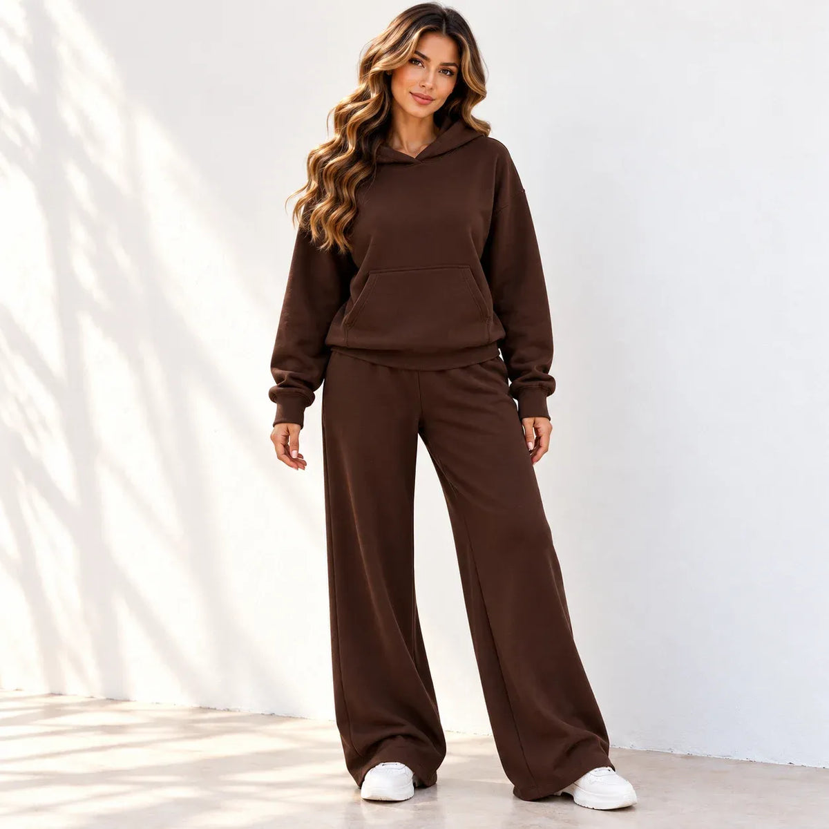 A woman modeling a matching brown two-piece loungewear set consisting of a pullover hoodie and wide-leg sweatpants, paired with white sneakers, standing against a white wall with soft leaf shadows.