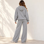 A woman wearing a light gray heathered two-piece loungewear set, consisting of a pullover hoodie and matching wide-leg sweatpants, shown from the back against a white wall with soft shadow patterns.