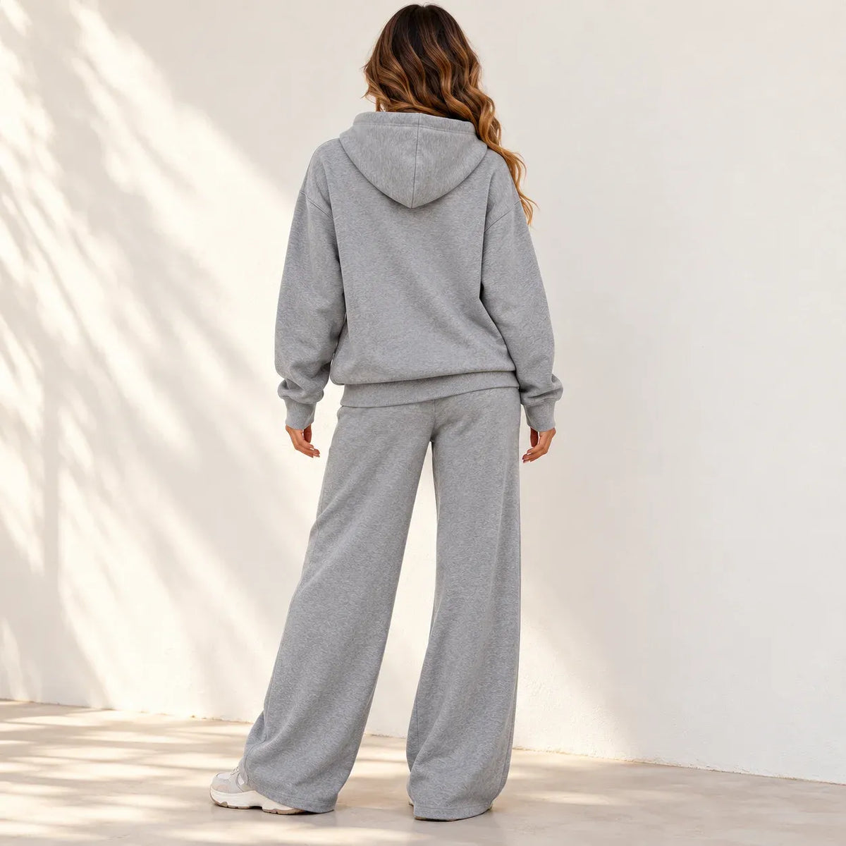A woman wearing a light gray heathered two-piece loungewear set, consisting of a pullover hoodie and matching wide-leg sweatpants, shown from the back against a white wall with soft shadow patterns.