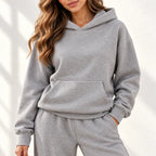 A woman modeling an grey two-piece loungewear set consisting of a pullover hoodie and matching wide-leg sweatpants, paired with white sneakers against a white wall with soft leaf shadows.