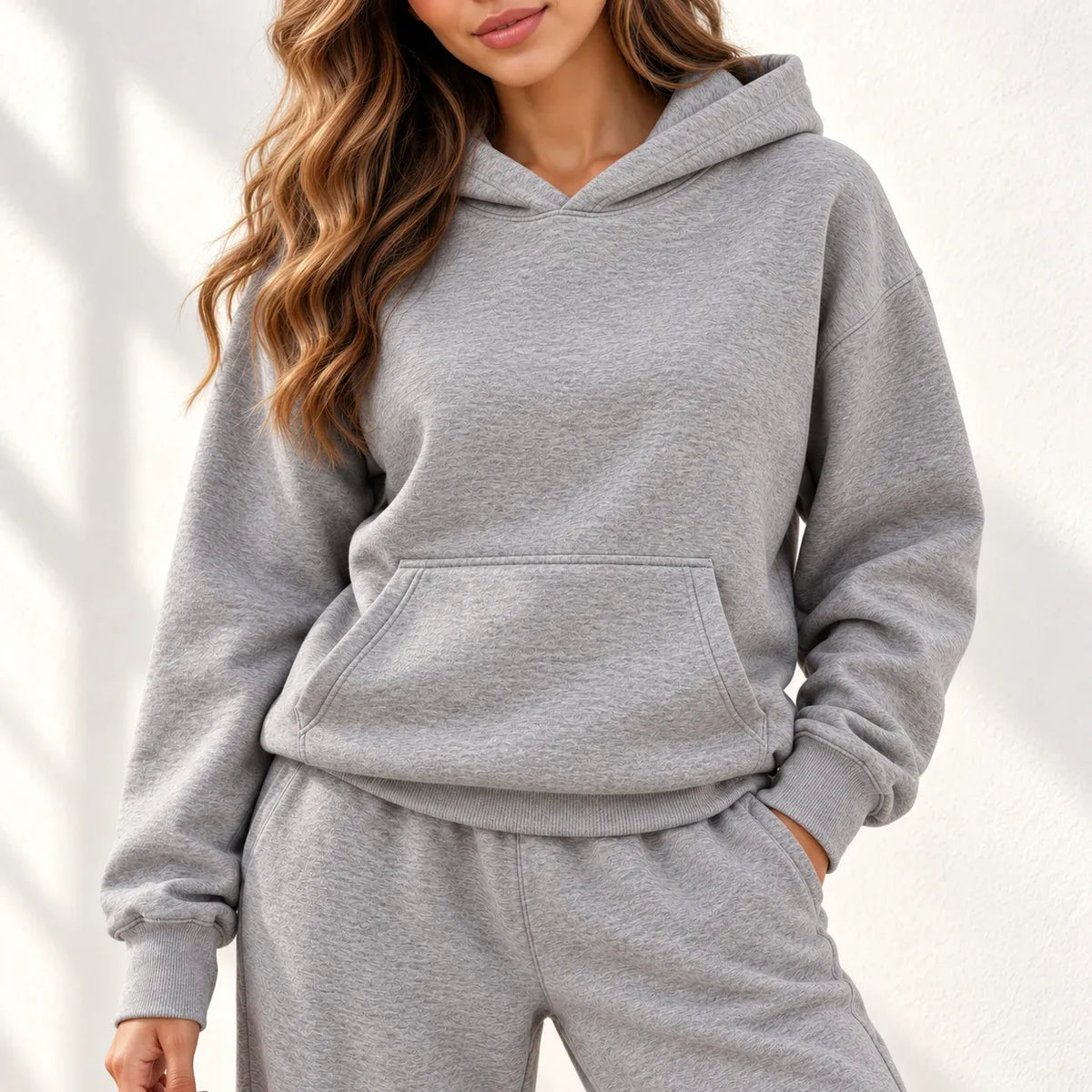 A woman modeling an grey two-piece loungewear set consisting of a pullover hoodie and matching wide-leg sweatpants, paired with white sneakers against a white wall with soft leaf shadows.