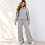 A woman modeling a light gray two-piece loungewear set consisting of a pullover hoodie and matching wide-leg sweatpants, standing against a white wall with soft shadows.