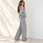 A woman wearing a matching light gray two-piece loungewear set, consisting of a pullover hoodie and wide-leg sweatpants, standing against a white wall with soft leaf shadows.