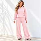A woman modeling an pink two-piece loungewear set consisting of a pullover hoodie and matching wide-leg sweatpants, paired with white sneakers against a white wall with soft leaf shadows.