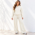 A woman modeling a matching cream-colored lounge set consisting of a pullover hoodie and wide-leg sweatpants, standing against a white wall with soft shadows.