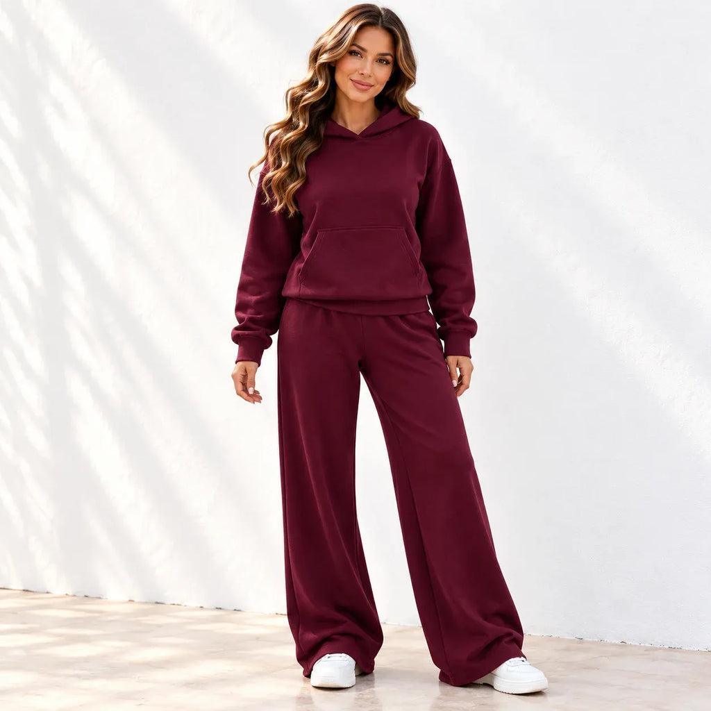 A woman modeling a matching burgundy lounge set consisting of a pullover hoodie and wide-leg sweatpants, paired with white sneakers, standing against a white wall with soft leaf shadows.