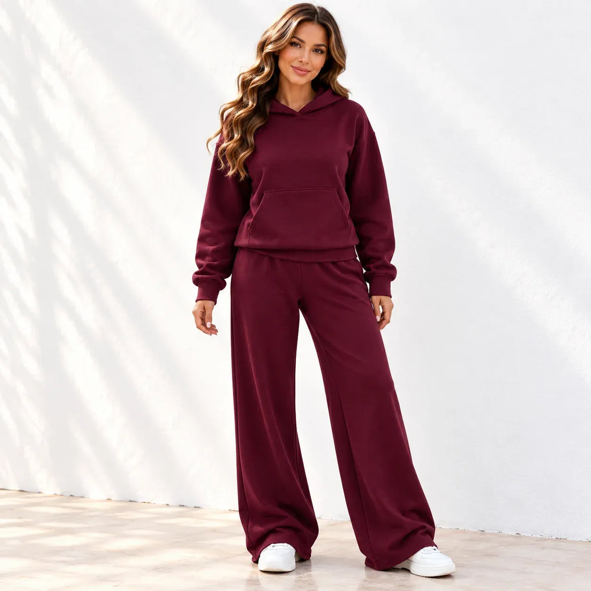 A woman modeling a matching burgundy lounge set consisting of a pullover hoodie and wide-leg sweatpants, paired with white sneakers, standing against a white wall with soft leaf shadows.