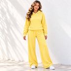 A woman modeling an yellow two-piece loungewear set consisting of a pullover hoodie and matching wide-leg sweatpants, paired with white sneakers against a white wall with soft leaf shadows.