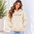 A woman wearing a beige pullover hoodie with the text 'you are enough.' printed in black across the chest, paired with light blue jeans, standing against a white wall with a potted plant on the left.