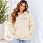 A woman wearing a beige pullover hoodie with the text 'you are enough.' printed in black across the chest, paired with light blue jeans, standing against a white wall with a potted plant on the left.