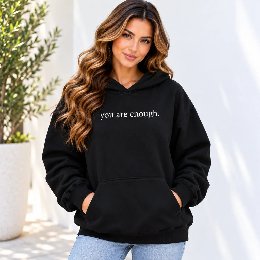 A woman wearing a black pullover hoodie with the phrase 'you are enough.' printed in white text across the chest, paired with light blue jeans, set against a bright, sunlit outdoor background with a potted plant.