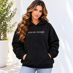 A woman wearing a black pullover hoodie with the phrase 'you are enough.' printed in white text across the chest, paired with light blue jeans, set against a bright, sunlit outdoor background with a potted plant.