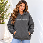 A woman wearing a dark gray heather pullover hoodie with the phrase 'you are enough.' printed in white lowercase serif text across the chest, paired with light blue jeans, standing against a white wall with a potted plant on the left.