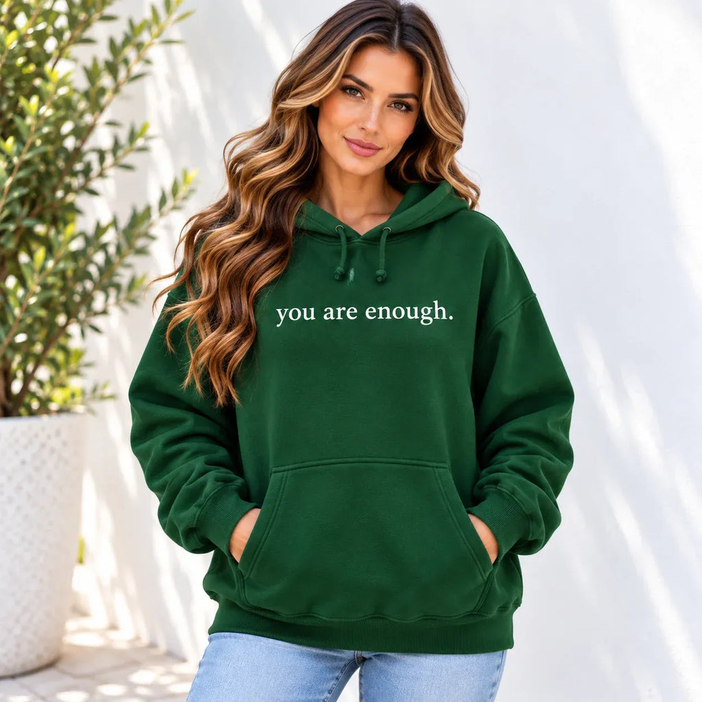 A woman wearing a forest green pullover hoodie with the phrase 'you are enough.' printed in white text across the chest, paired with light blue jeans, set against a bright white wall with soft shadows.
