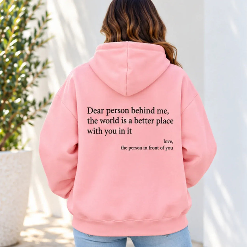 A woman viewed from behind wearing a light pink hoodie with black printed text on the back that reads, 'Dear person behind me, the world is a better place with you in it. love, the person in front of you.