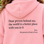 A close-up of a person wearing a light pink hoodie, showing the back of the garment with black printed text that reads, 'Dear person behind me, the world is a better place with you in it love, the person in front of you'.