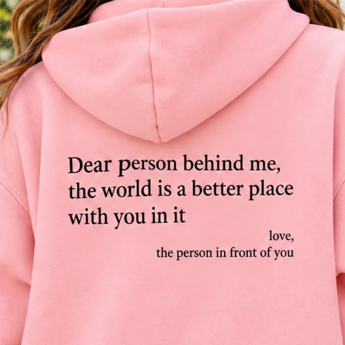 A close-up of a person wearing a light pink hoodie, showing the back of the garment with black printed text that reads, 'Dear person behind me, the world is a better place with you in it love, the person in front of you'.