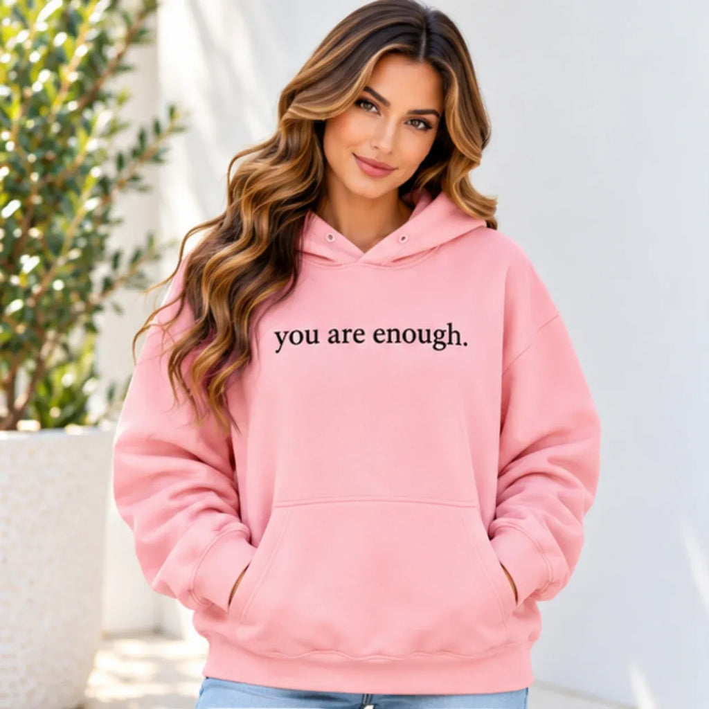 A woman wearing a light pink pullover hoodie with the text 'you are enough.' printed in black across the chest, posed outdoors against a light wall with a plant in the background.