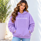 A woman wearing a lavender purple hoodie with the text 'you are enough.' printed in white across the chest, paired with light blue jeans, standing against a white wall with a potted plant on the left.