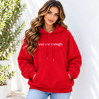 A woman wearing a bright red pullover hoodie with the phrase 'you are enough.' printed in white across the chest, paired with light blue jeans, standing against a white wall with potted plants in the background.