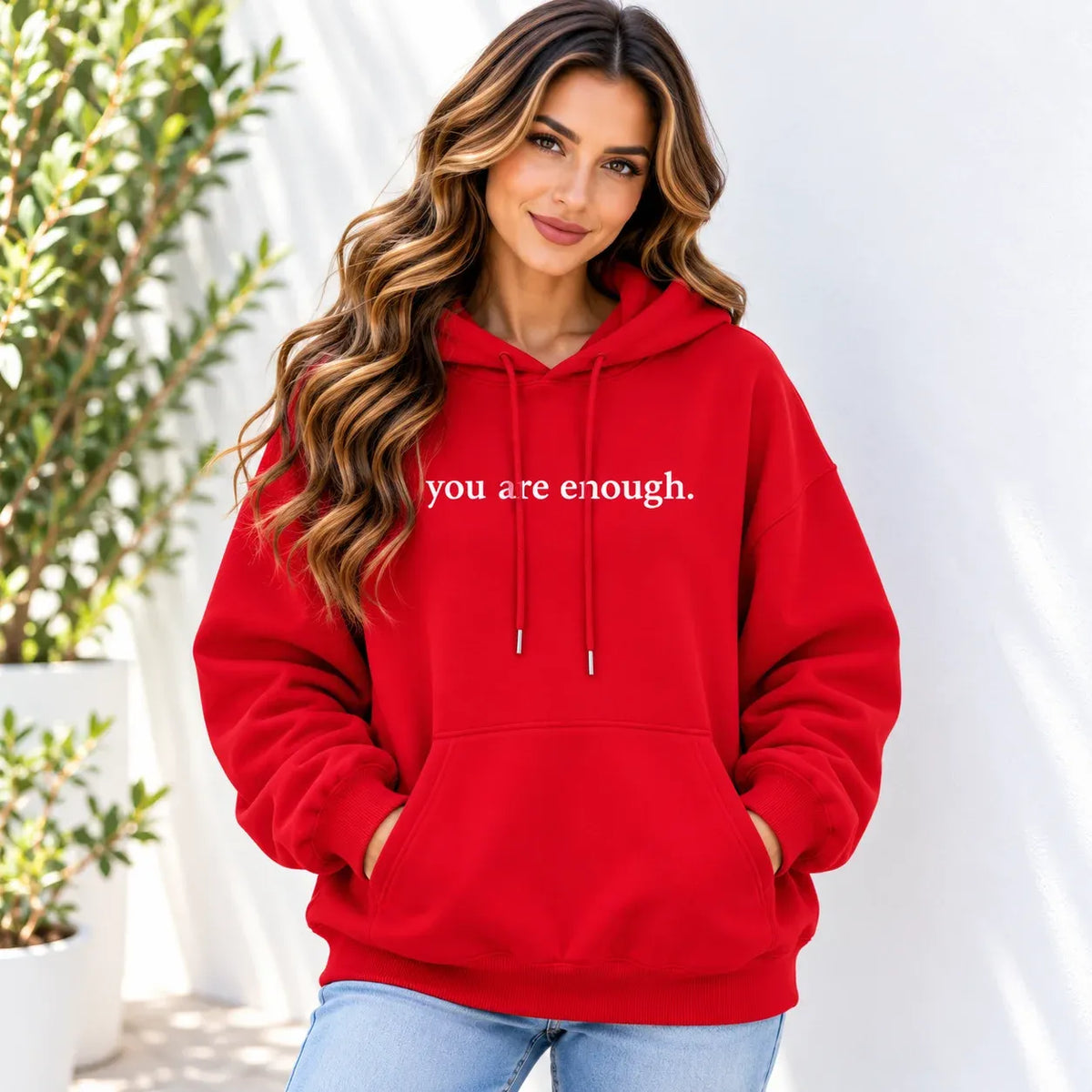 A woman wearing a bright red pullover hoodie with the phrase 'you are enough.' printed in white across the chest, paired with light blue jeans, standing against a white wall with potted plants in the background.
