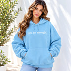 A woman wearing a light blue pullover hoodie with the phrase 'you are enough.' printed in white across the chest, paired with light-wash jeans, set against a bright white wall with soft shadows and green plants on the side.