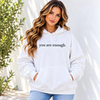 A woman wearing a white pullover hoodie with the text 'you are enough.' printed in black lowercase serif font across the chest, paired with light blue jeans, standing against a white wall with soft shadows.