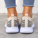 A pair of beige slip-on sneakers with black knit stripe patterns on the heel and thick white soles, shown on a person's feet wearing blue jeans.