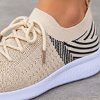 Close-up of a beige knit slip-on sneaker with cream laces, a black and beige patterned heel, and a white sole, shown on a person's foot against a gray pavement background.