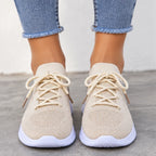 A pair of beige knit lace-up sneakers worn with frayed blue jeans, shown from the front against a neutral concrete and wall background.