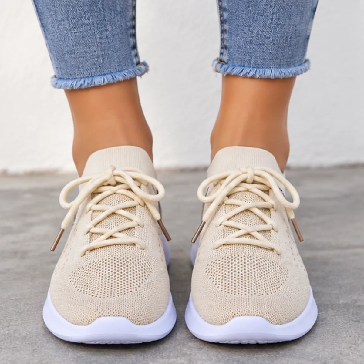 A pair of beige knit lace-up sneakers worn with frayed blue jeans, shown from the front against a neutral concrete and wall background.