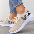 A close-up of a person wearing beige knit slip-on sneakers with black striped knit panels and white soles, paired with light blue frayed-hem jeans, standing on a concrete floor against a light wall.