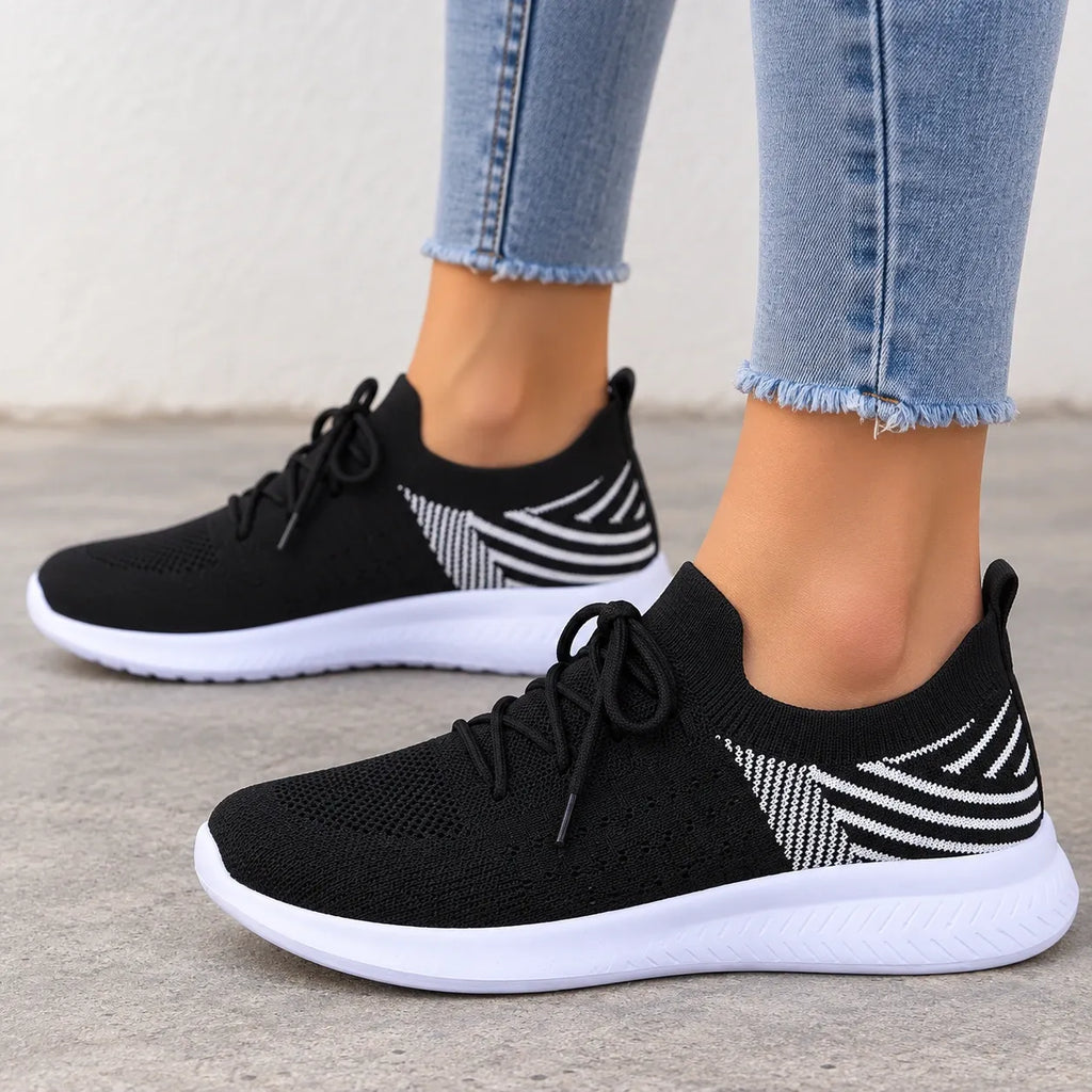 A pair of black knit slip-on sneakers with white soles and white geometric side patterns, shown on a model wearing light blue frayed-hem jeans against a concrete floor and wall.
