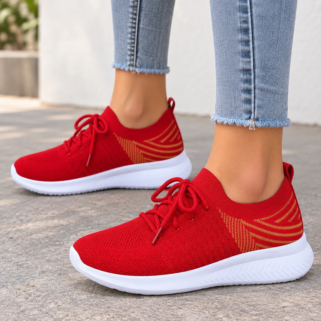 A pair of red knit slip-on sneakers with white soles and gold geometric side accents, shown on a person wearing light blue frayed-hem jeans outdoors on a paved surface.