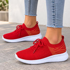 A pair of red knit slip-on sneakers with white soles and gold geometric side accents, shown on a person wearing light blue frayed-hem jeans outdoors on a paved surface.