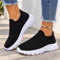 A pair of women's black knit slip-on sneakers with chunky white platform soles, shown being worn with frayed blue jeans and a beaded ankle bracelet.