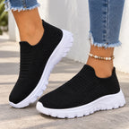 A pair of women's black knit slip-on sneakers with chunky white platform soles, shown being worn with frayed blue jeans and a beaded ankle bracelet.