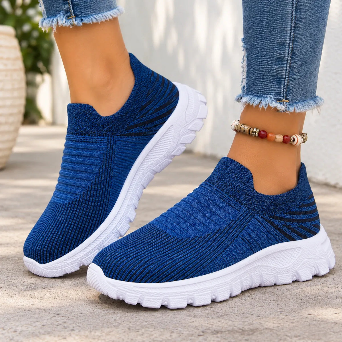 A pair of women's slip-on knit sneakers in a deep blue color with chunky white soles, shown being worn with frayed blue jeans.