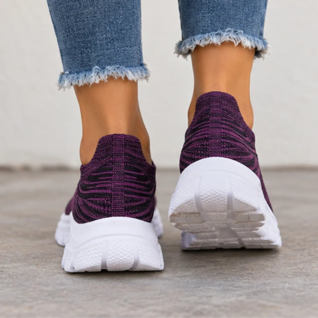 A pair of slip-on knit sneakers in deep purple and white, worn with frayed blue jeans on a concrete floor.