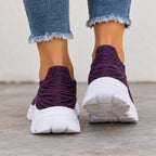 A pair of slip-on knit sneakers in deep purple and white, worn with frayed blue jeans on a concrete floor.