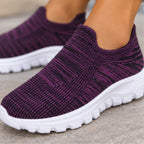 A close-up of a person wearing slip-on knit sneakers with a deep purple and black textured upper and chunky white soles, standing on a gray concrete surface.