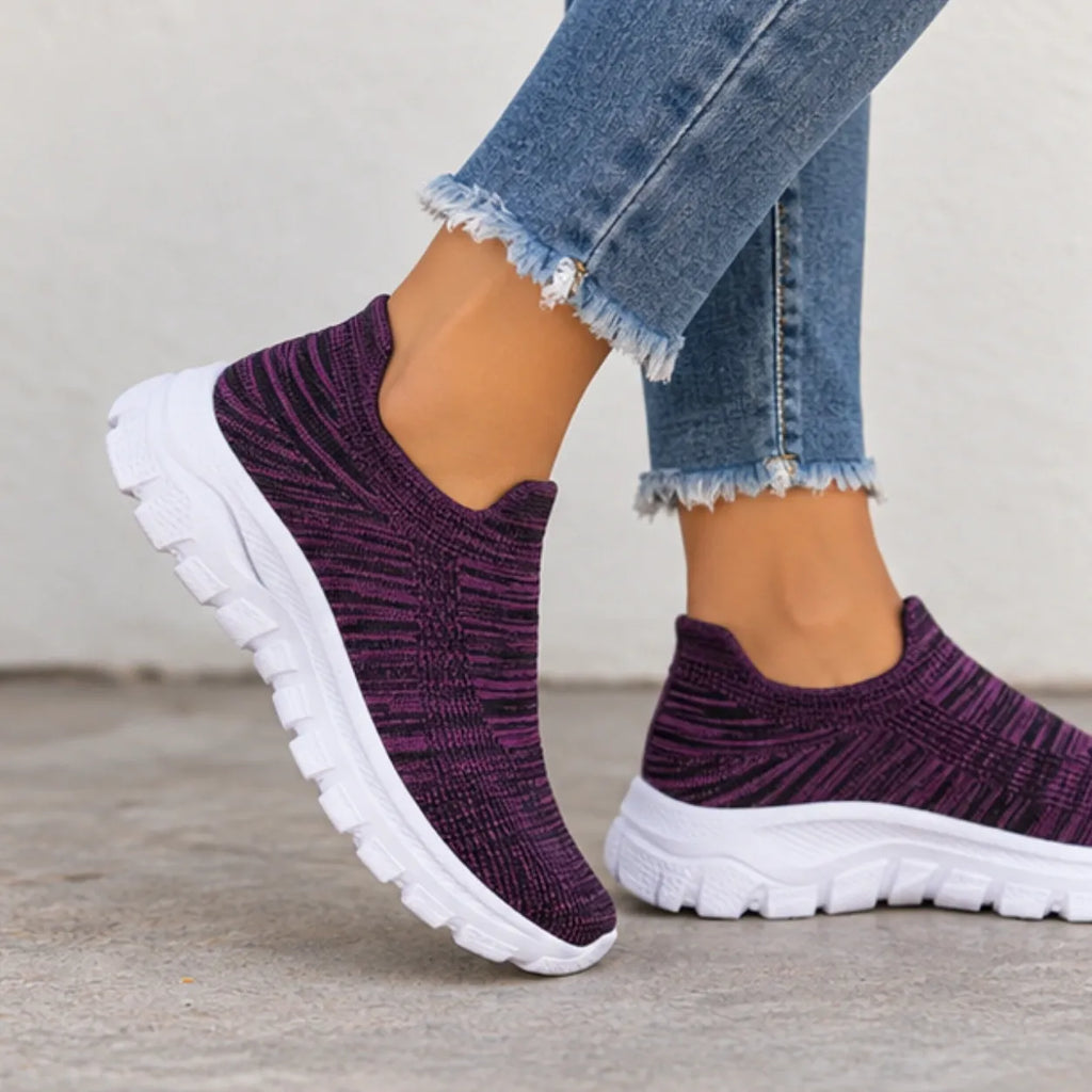 A person wearing slip-on knit sneakers in a deep purple and black pattern with chunky white soles, paired with frayed blue jeans.