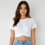 A woman wearing a white short-sleeve crop top with a twisted knot detail at the front, paired with light blue jeans, against a plain light gray background.