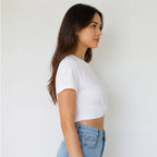 A woman in a side profile wearing a white short-sleeve crop top and light blue jeans, standing against a plain white wall.