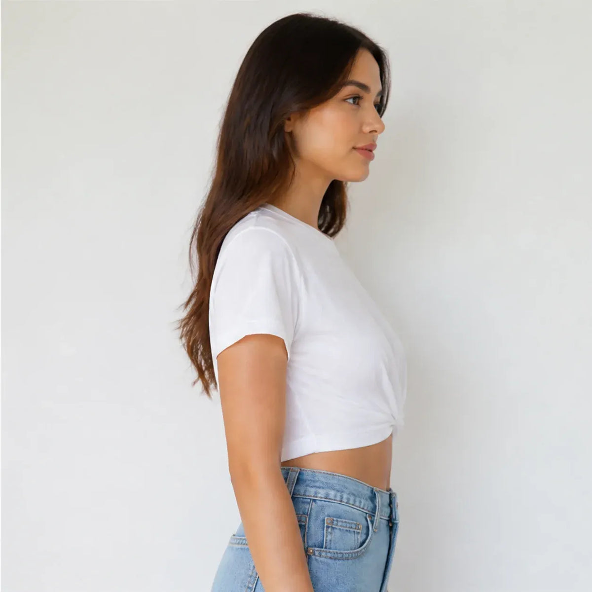 A woman in a side profile wearing a white short-sleeve crop top and light blue jeans, standing against a plain white wall.